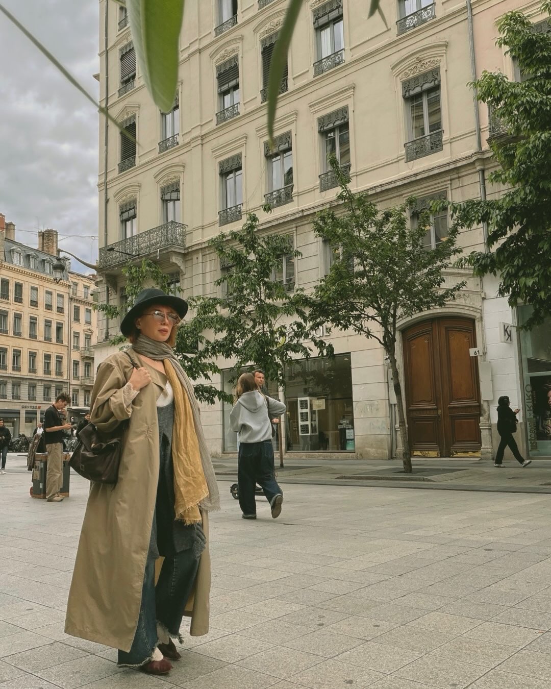 Jasmine Petrosyan styled in a long trench coat and layered scarves on a Lyon street