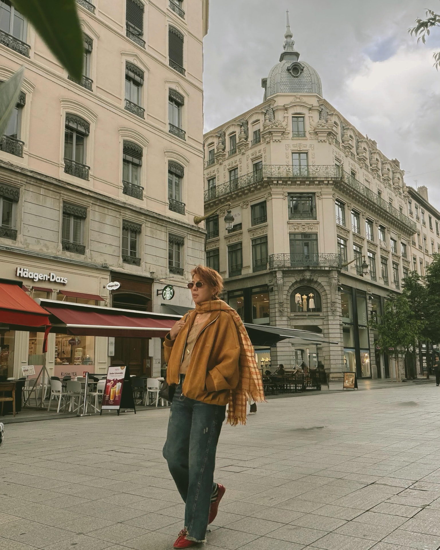 Jasmine Petrosyan in an amber suede jacket and checked scarf in a European city square