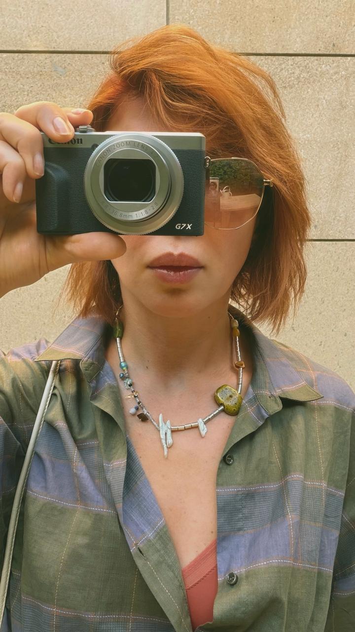 Jasmine holding a compact camera, wearing a checked shirt and a stone bead necklace