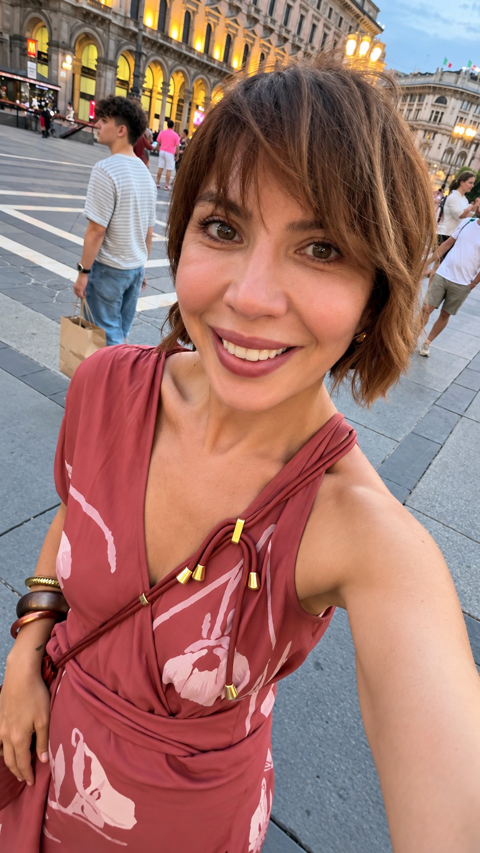 Jasmine Petrosyan in a terracotta floral wrap dress with gold-tipped cords on a Milan piazza at dusk