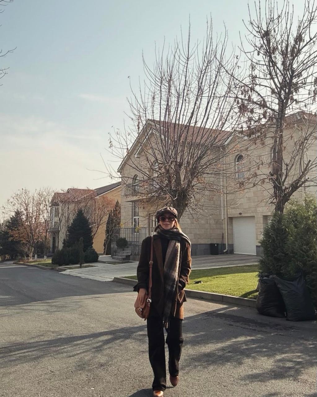 Jasmine in a dark coat, plaid scarf and baker boy cap on a sunlit Yerevan street